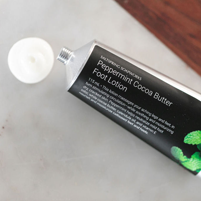 A squeeze tube of Saltspring Soapworks Peppermint Cocoa Butter Foot Lotion lies on a light marble surface, with a dark wood surface in the background.