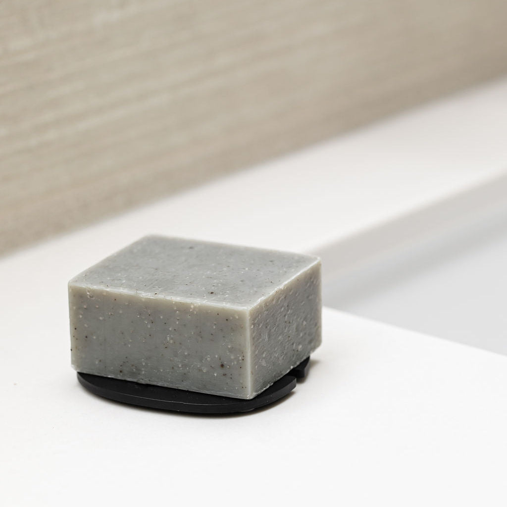 A rectangular bar of gray soap with visible speckles, resting on a black Saltspring Soapworks Soap Lift on a white surface with a beige textured background.