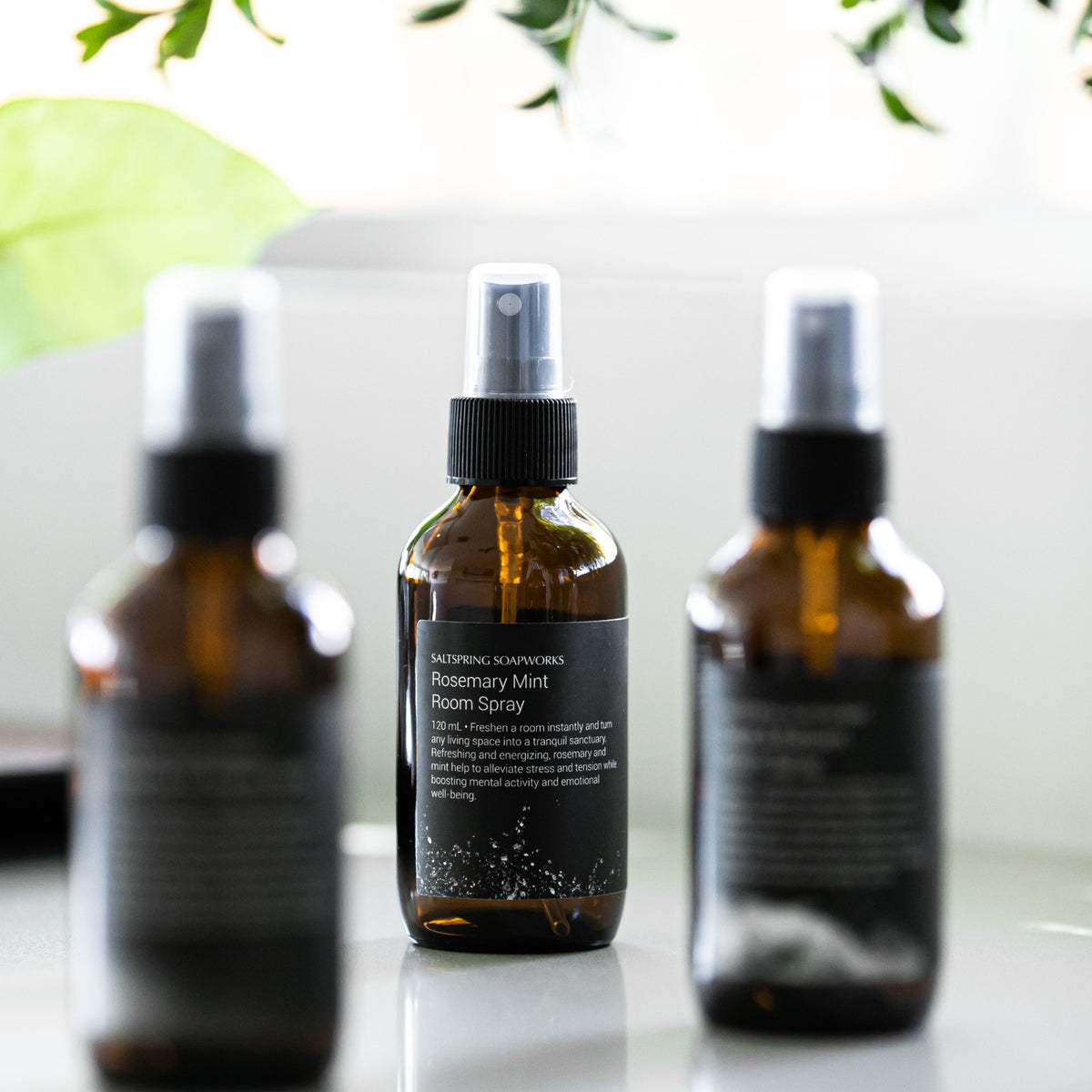 Three amber glass spray bottles with black caps and black labels are arranged on a glossy white surface. The center bottle is in sharp focus, showing the label text 'Rosemary Mint Room Spray' while the two bottles on the sides are softly blurred.
