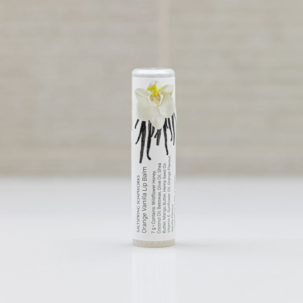 A single tube Saltspring Soapworks Orange Vanilla lip balm standing upright on a white surface against a soft beige textured background. The tube is white with black text and a white orchid type flower illustration with a small yellow center.