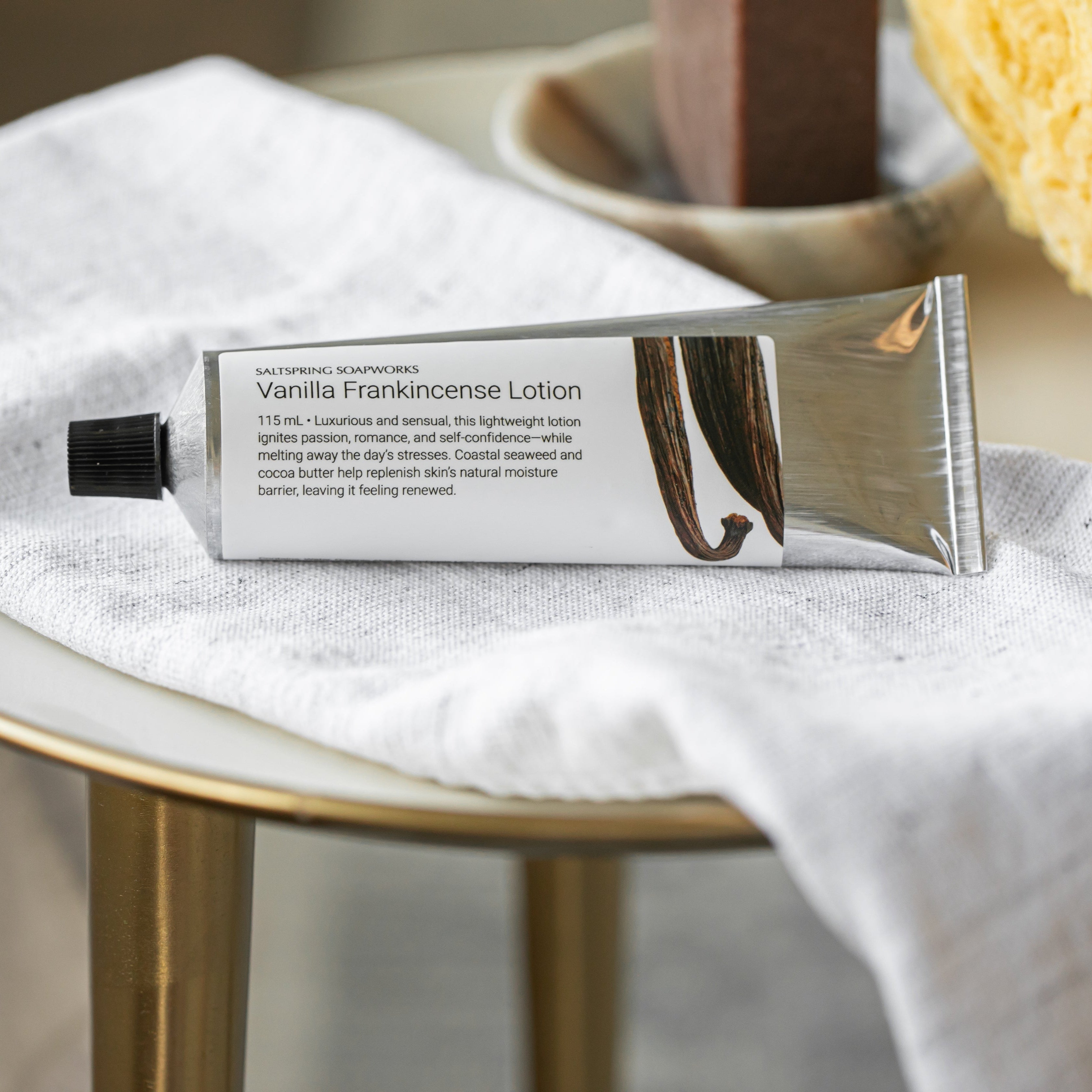 Vanilla Frankincense Lotion tube on a white cloth with a sponge and soap dish in the background.