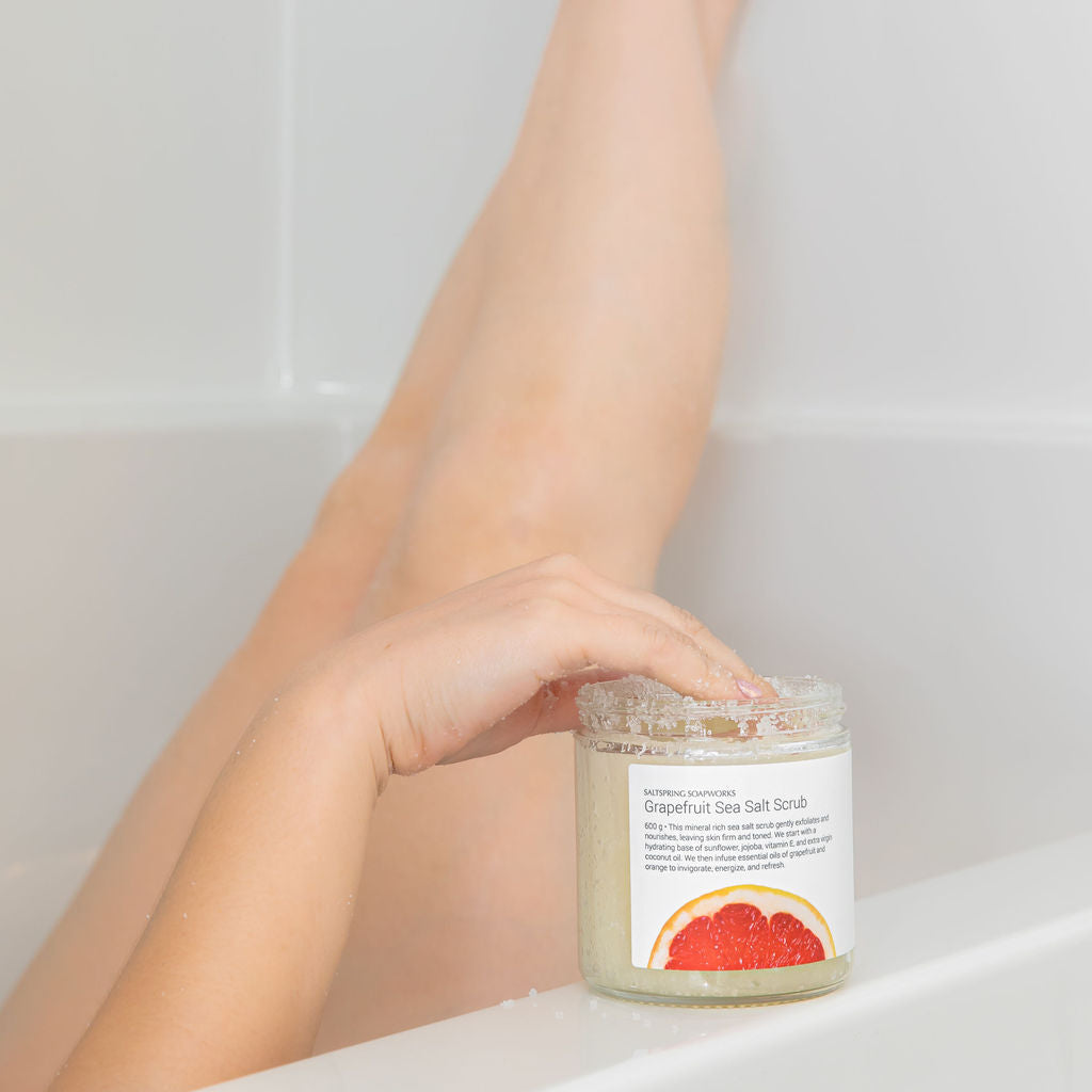 Person holding a jar of exfoliating foot scrub with their feet in the background