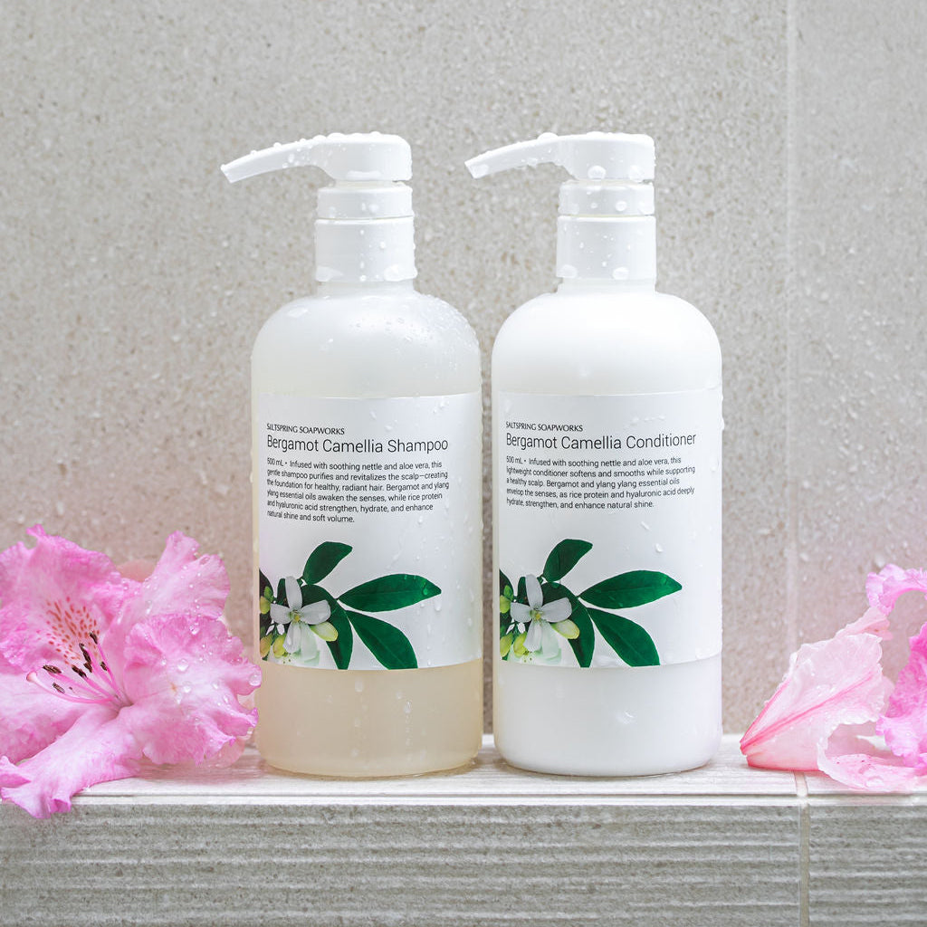 Two bottles of Saltspring Soapworks Bergamot Camellia shampoo and conditioner with floral blossoms on a tiled surface.