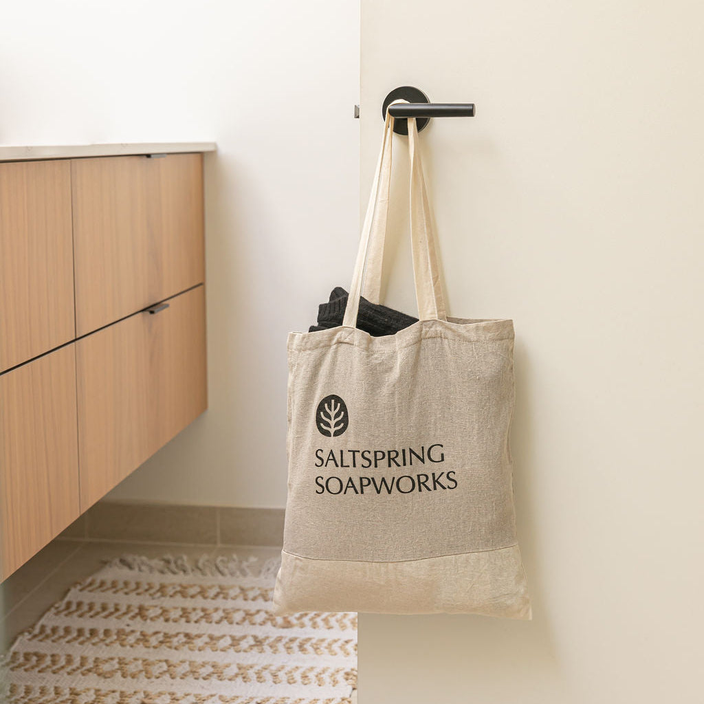 A beige canvas tote bag with black text reading 'SALTSPRING SOAPWORKS' hanging from a black door knob in a minimalist bathroom setting.