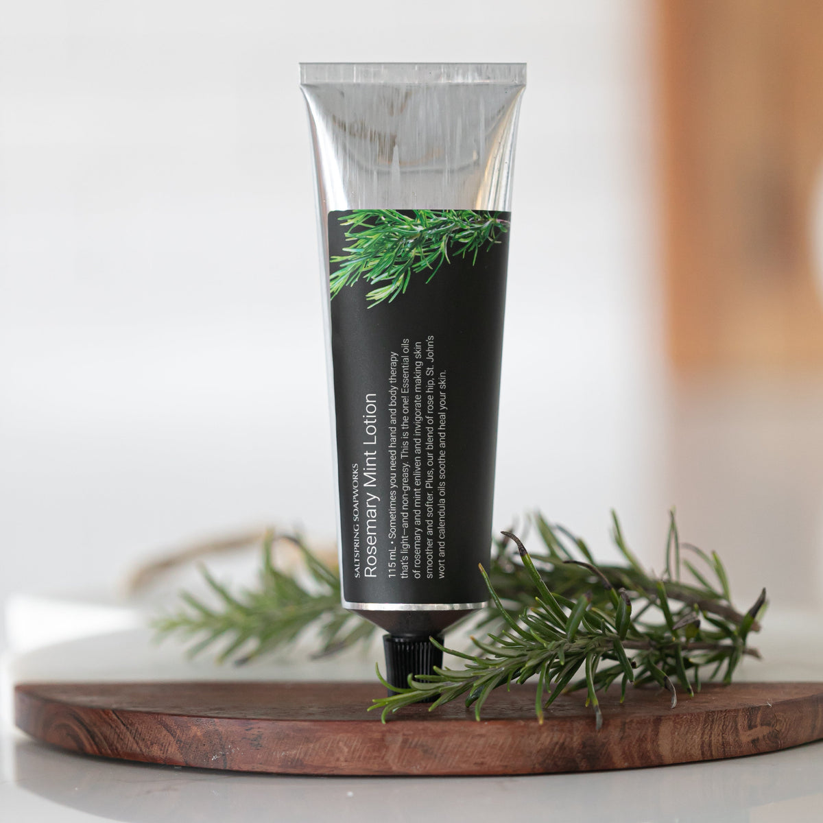 Saltspring Soapworks Rosemary Mint body lotion stands upright on a round wooden board with rosemary sprigs around it. 