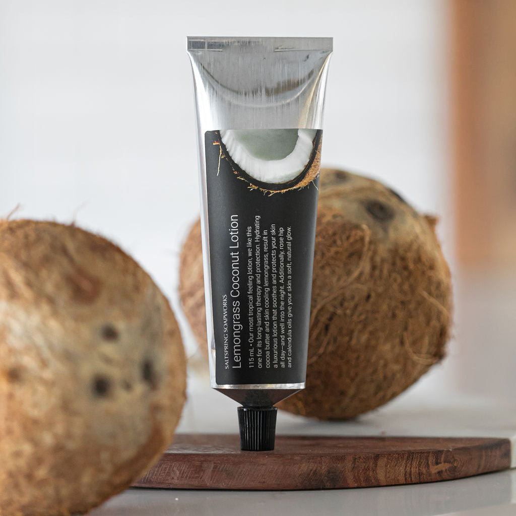A squeeze tube of Saltspring Soapworks Lemongrass Coconut Lotion stands upright on a small wooden board, with two whole coconuts blurred in the foreground and background on a glossy white surface.