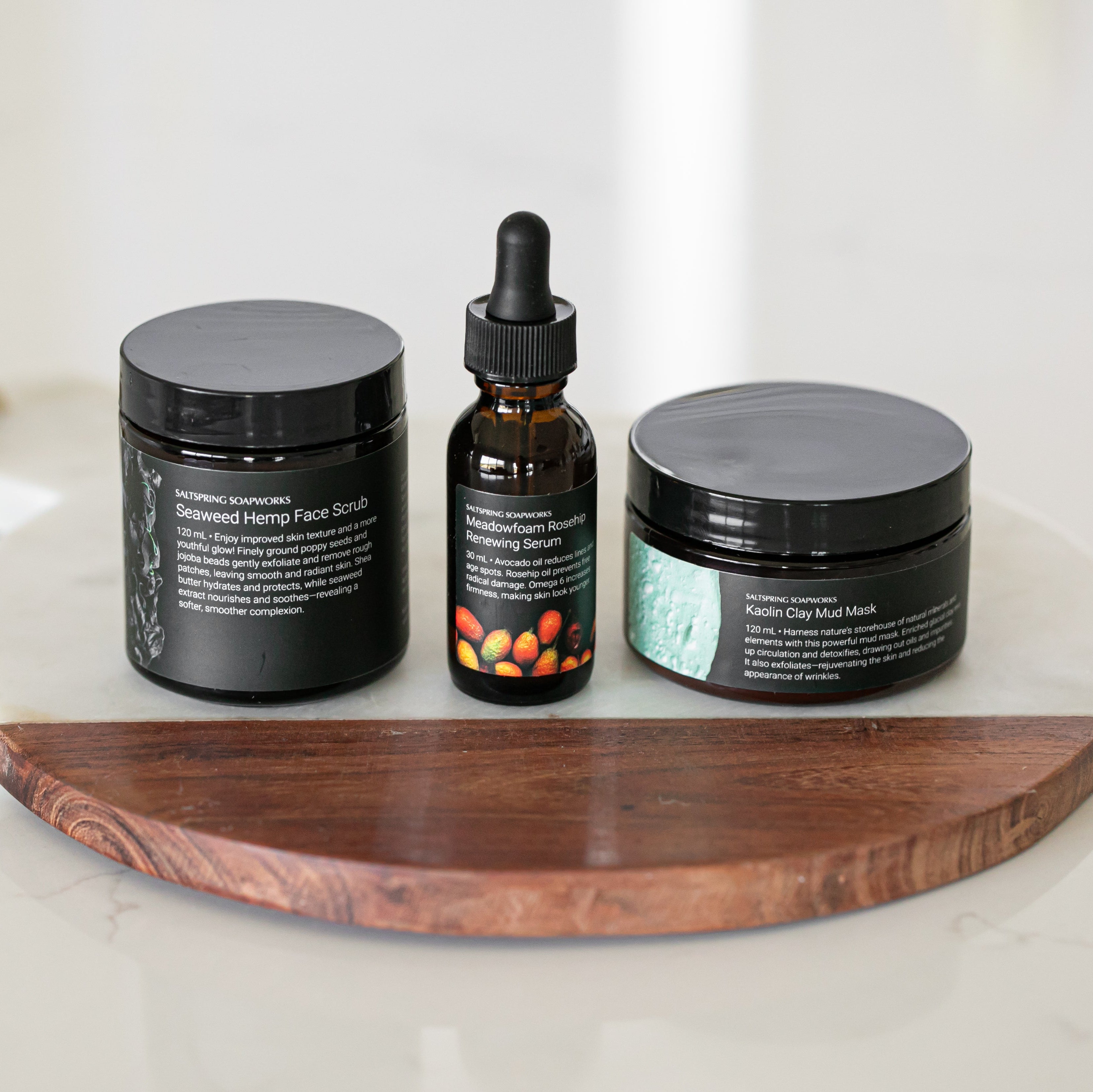 Three skincare products, a seaweed hemp scrub exfoliator, a face oil serum and a Kaolin clay mud mask on a wooden tray with a white background