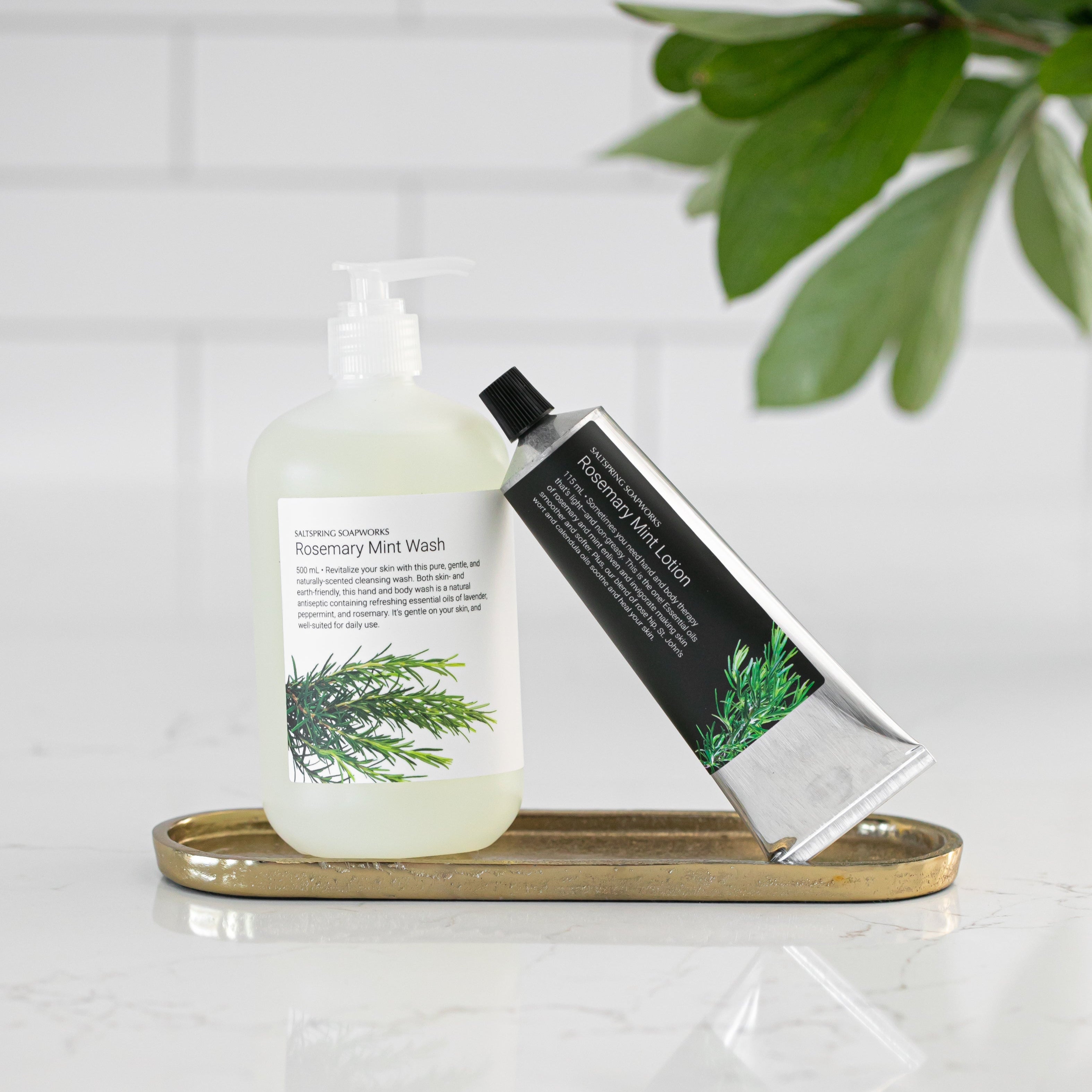 Bottle of Rosemary Mint liquid soap and a lotion leaning against it, on a brass tray with a plant in the background
