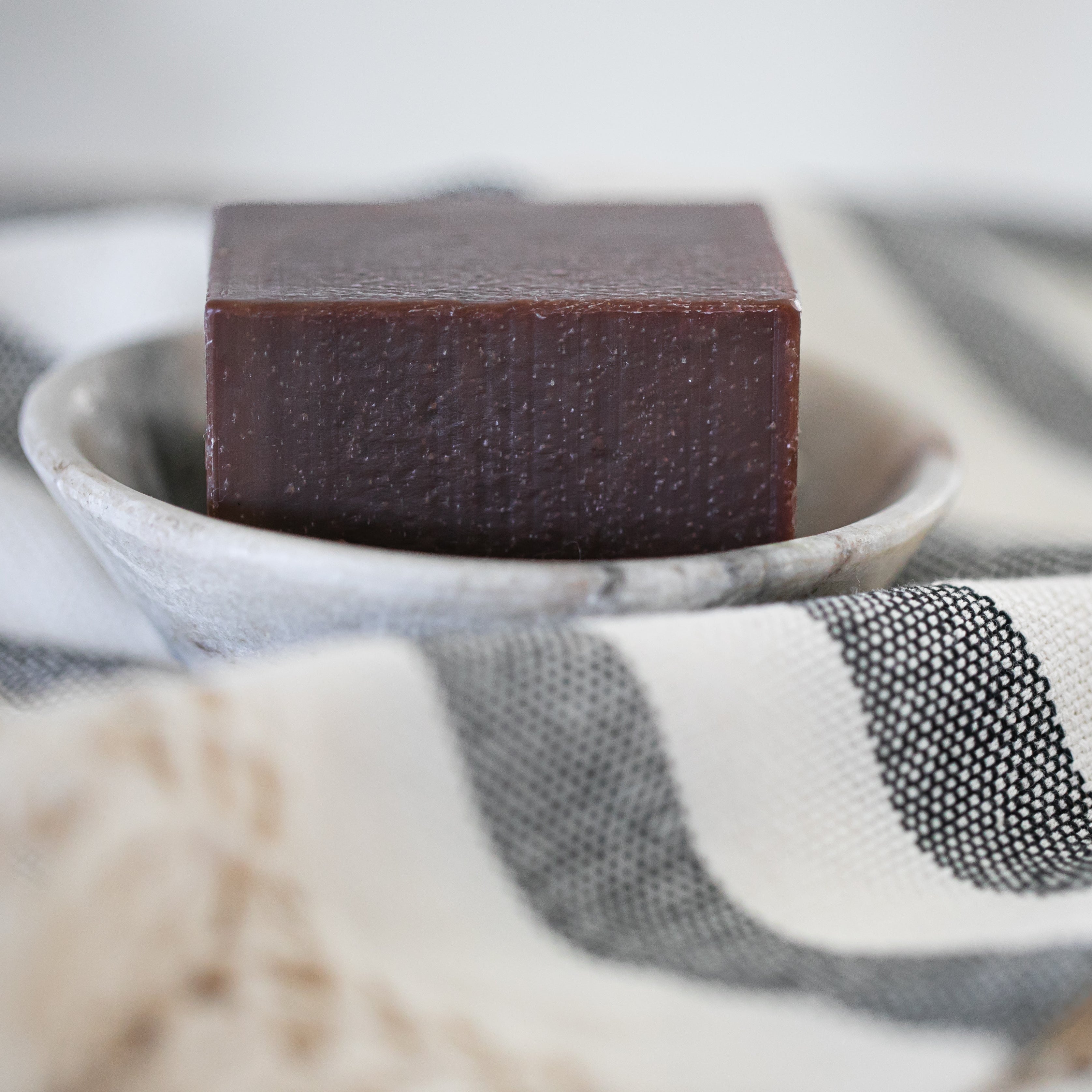 Rich brown bar of soap in a stone bowl with a striped towel in the background