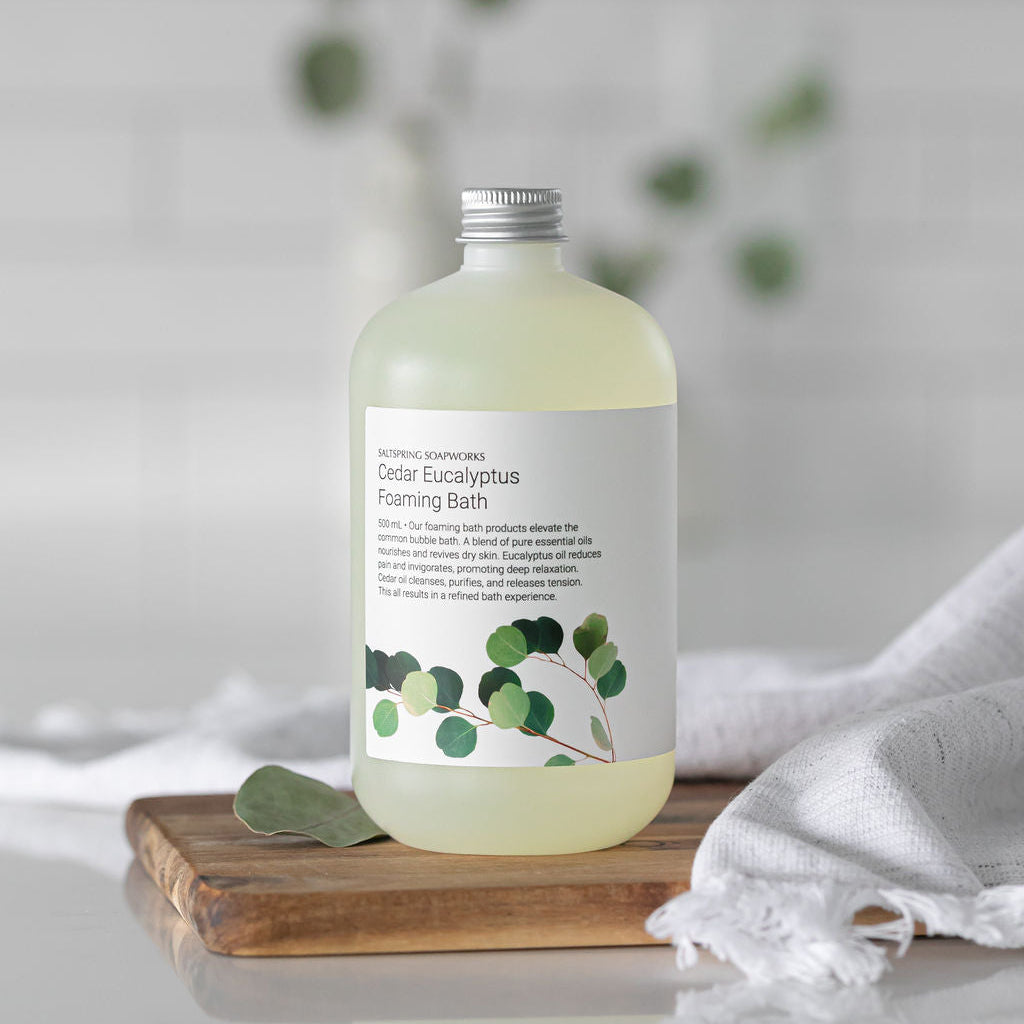 Saltspring Soapworks Cedar Eucalyptus Foaming Bath on a wooden board with a linen towel and green leaves in the background.