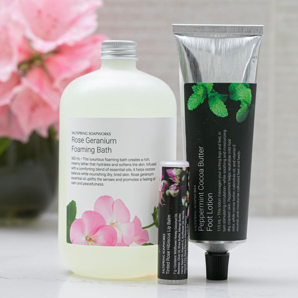 Rose Geranium Foaming Bath, Peppermint Cocoa Butter Foot Lotion, and Tinted Rose Hibiscus Lip Balm on a light surface with pink flowers in the background.