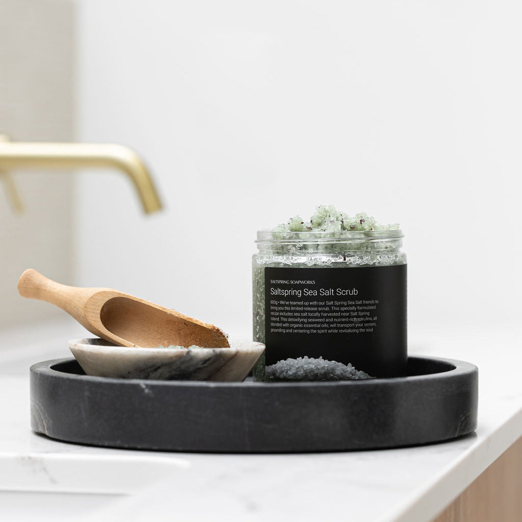 Saltspring Sea Salt Scrub open jar, a wooden scoop, on a black tray on a bathroom counter.