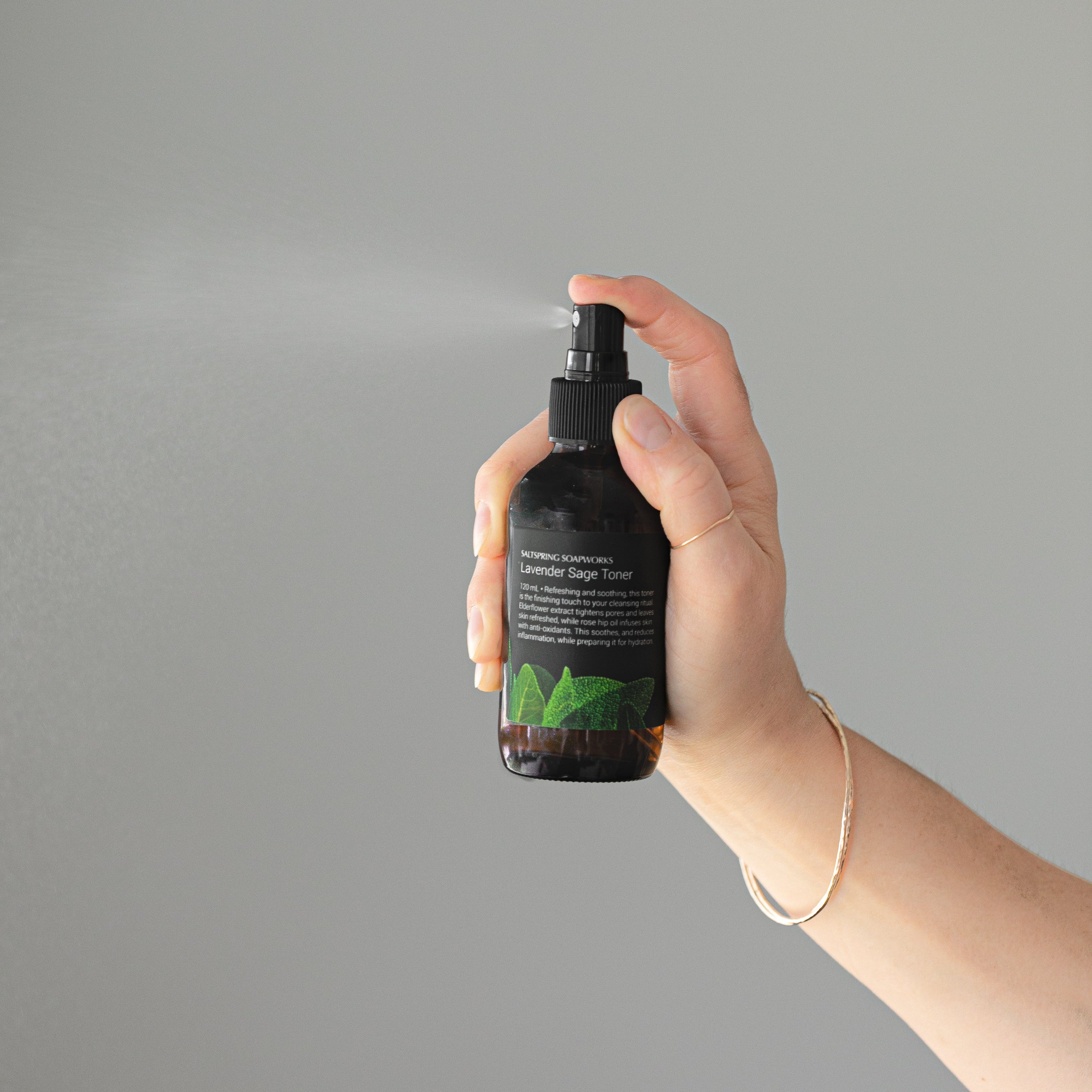Hand holding a black spray bottle with a lavender sage toner label against a gray background